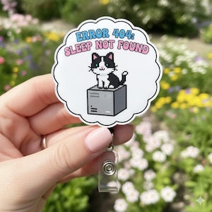 Può includere: Un badge bianco con un gatto pixelato in bianco e nero su una scatola grigia, con la scritta "ERROR 404: SLEEP NOT FOUND" in blu e rosa. Il badge ha un avvolgitore retrattile in plastica trasparente.