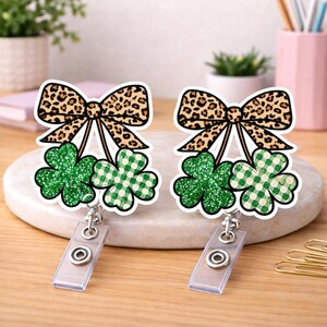 Puede incluir: Dos portatarjetas retráctiles con temática del Día de San Patricio. Cada uno presenta un lazo con estampado de leopardo, un trébol con purpurina verde y un trébol a cuadros verde y blanco. Los portatarjetas tienen una correa de plástico transparente y un clip metálico.