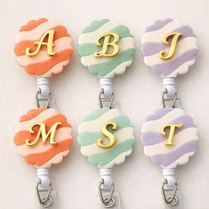 May include: Six colourful badge reels with gold letters A, B, J, M, S, and T. Each reel features a scalloped edge and a striped design in orange, green, and purple. The reels have a white retractable cord and a metal clip.