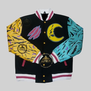 Pode incluir: Casaco universitário preto com detalhes em amarelo, azul e roxo. As mangas apresentam desenhos de listras de tigre e abstratos. Uma lua crescente amarela e outros patches adornam a frente. O casaco tem gola e punhos brancos com listras vermelhas. O casaco tem o texto "Apex Custom".