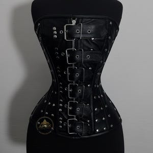 以下が含まれることがあります： シルバーのリベットとバックルが付いた黒い革のコルセット。コルセットはジッパー開閉と複数の調節可能なストラップを備えています。左下にロゴが見えます。コルセットは黒いマネキンに表示されています。