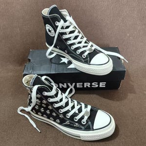 Puede incluir: Zapatillas altas negras con cordones blancos y puntera blanca. Las zapatillas tienen un logotipo de estrella y tachuelas plateadas. Están sobre una caja negra con la palabra "CONVERSE".