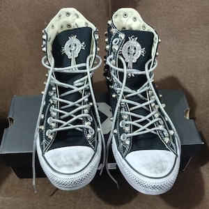 Puede incluir: Par de zapatillas altas negras con tachuelas y ojales de metal plateado. Las zapatillas tienen cordones blancos y una puntera de goma blanca. Cada zapato presenta un emblema plateado en la lengüeta. Los zapatos están encima de una caja de zapatos negra.