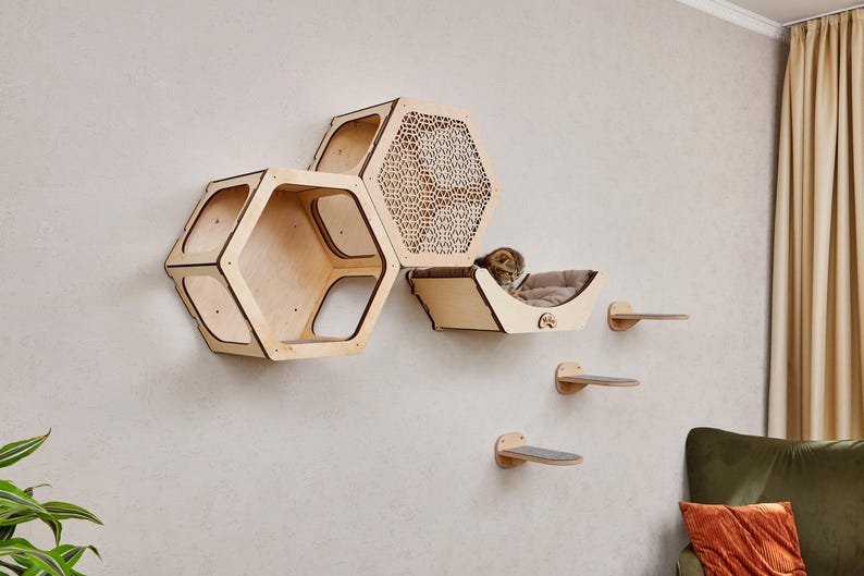 Puede incluir: Un juego de muebles para gatos montado en la pared con acabado de madera natural. El juego incluye tres estantes hexagonales, una cama para gatos y tres peque&ntilde;os escalones. Un gato descansa en la cama. Los muebles est&aacute;n montados en una pared gris claro.