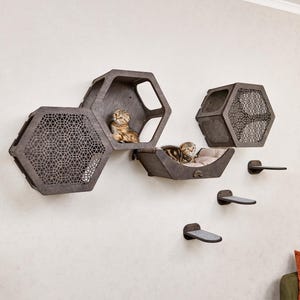Puede incluir: Un conjunto de muebles para gatos montado en la pared con un dise&ntilde;o moderno. El conjunto incluye tres casas hexagonales para gatos y tres estantes escalonados, todos en un acabado gris oscuro. Dos gatos descansan dentro de las casas.