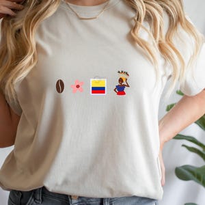 Puede incluir: Camiseta color crema con un diseño que incluye un grano de café, una flor rosa, la bandera colombiana y una mujer con una cesta en la cabeza. El diseño está centrado en la parte delantera.