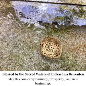 Op de afbeelding: Een geweven mandje gevuld met Japanse munten ligt op een getextureerd stenen oppervlak bij stromend water. De afbeelding bevat de tekst "Blessed by the Sacred Waters of Inokashira Benzaiten" en een boodschap over harmonie en voorspoed.