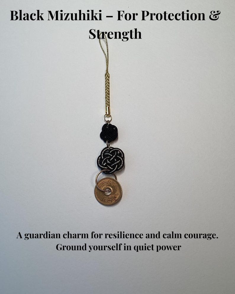 Op de afbeelding: Een zwarte Mizuhiki-bedel met een gouden koord, met twee decoratieve knopen en een Japanse munt. De tekst luidt "Black Mizuhiki - For Protection & Strength" en "A guardian charm for resilience and calm courage."