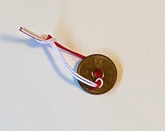 Op de afbeelding: Een bronzen Japanse munt met een centraal gat, bevestigd aan een rood-wit koord. De munt is rond met een vierkant ontwerp. Het koord is bovenaan geknoopt.