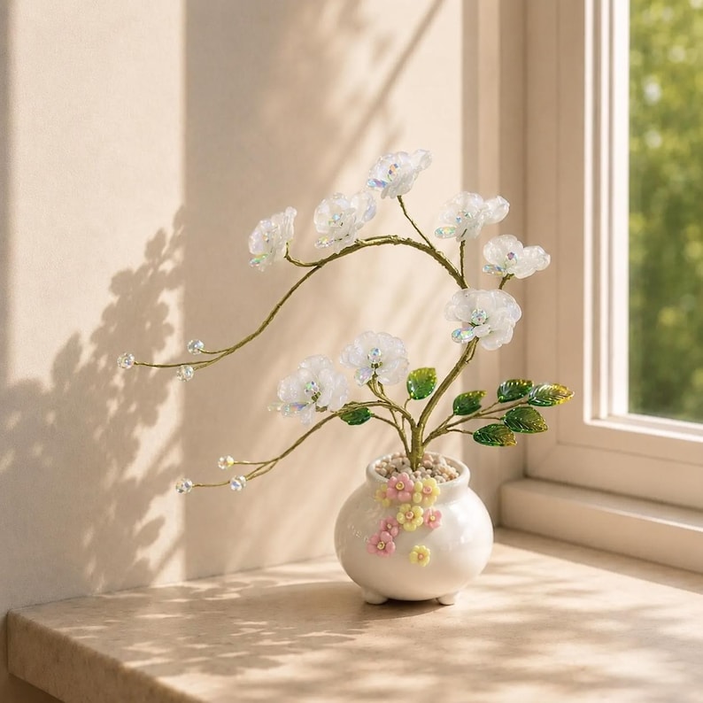May include: A decorative sculpture of a flowering tree with translucent white petals and green leaves on thin gold wire branches. The tree is set in a small, round white ceramic pot adorned with tiny pink and yellow floral accents.