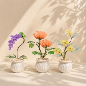 May include: Three decorative potted plants featuring translucent purple, orange, and yellow floral elements on thin wire stems with green leaves, each set in a small white ceramic pot filled with light-coloured pebbles for a display.