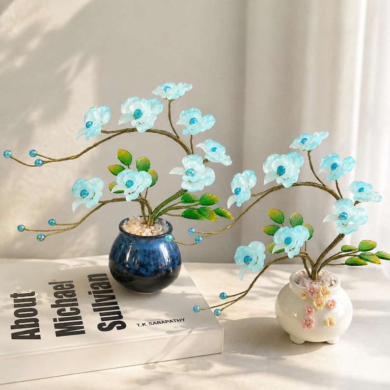 May include: Two decorative bonsai-style ornaments featuring translucent light blue flowers with bright blue centers, green leaves, and thin gold-toned wire branches. One sits in a dark blue pot, the other in a white pot with small pink floral accents.