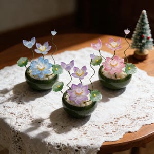 Puede incluir: Tres arreglos decorativos en miniatura de flores de loto en pequeñas macetas de cerámica verde. Cada arreglo presenta una flor de loto de diferente color, incluyendo azul, rosa y morado, con hojas verdes y guijarros blancos.