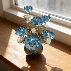 Può includere: Una scultura decorativa composta da diversi fiori di loto blu traslucidi con steli e centri dorati, disposti in un piccolo vaso in ceramica rotondo di colore scuro con una finitura maculata, creando una composizione floreale variegata.