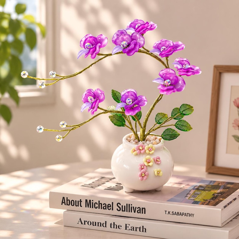 May include: A decorative arrangement featuring vibrant purple flowers with central iridescent crystals, set on thin gold-toned wire stems with small clear beads. The flowers are planted in a small white ceramic pot adorned with tiny pastel floral accents.
