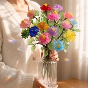 Op de afbeelding: Een heldere glazen vaas bevat een levendig boeket van kleurrijke kunstbloemen. De bloemen zijn in tinten rood, roze, oranje, geel, blauw en paars, met groene bladeren. De vaas heeft een geribbeld ontwerp.