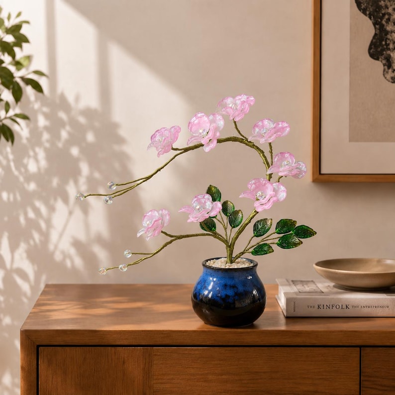 May include: A decorative artificial plant featuring translucent light pink flowers with clear crystal centers, green leaves, and thin wire stems, potted in a small, round, glossy blue and black ceramic vase filled with white pebbles.