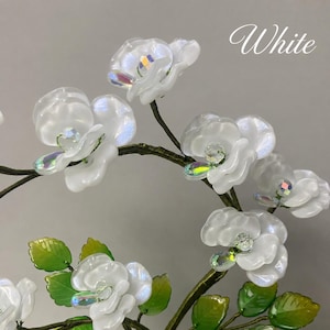 May include: A decorative branch featuring translucent white glass flowers with iridescent centers and green glass leaves attached to a dark brown wire stem. The word White is printed in a cursive script in the upper right corner of the image.