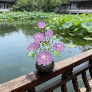 May include: A decorative ornament featuring multiple translucent purple lotus flowers and small green lily pads attached to thin, winding gold-toned metal stems, all set within a small, round, dark speckled ceramic vase.