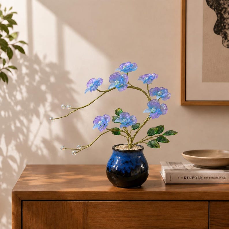 May include: A decorative artificial orchid plant with translucent blue and purple petals, green leaves, and thin stems tipped with small clear beads. The plant is potted in a small, round, dark blue ceramic vase filled with light-colored pebbles.
