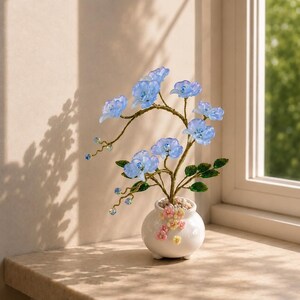 May include: A decorative sculpture of a flowering plant with translucent light blue petals and green leaves on gold-toned wire branches. The plant is set in a small, round white ceramic pot adorned with tiny pink and yellow floral accents.