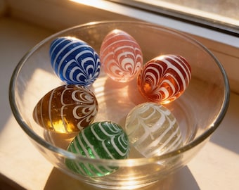 May include: A clear glass bowl filled with seven decorative glass eggs. The eggs are various colors, including blue, red, green, and amber, with white swirled patterns. The bowl sits near a window, illuminated by sunlight.