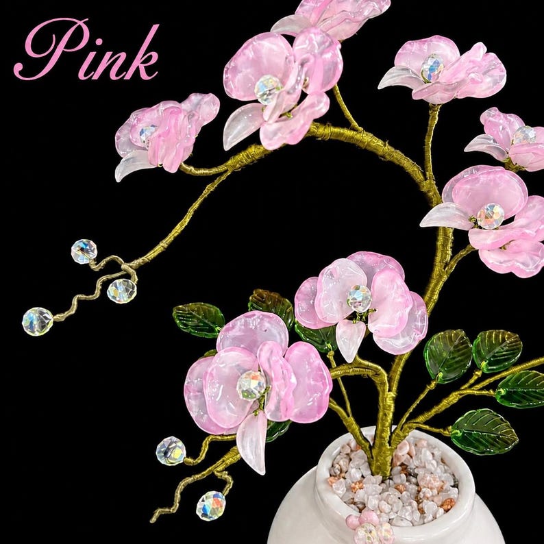 May include: A decorative tree sculpture featuring translucent pink flower petals with iridescent crystal centers, green glass leaves, and gold-toned wire branches. The tree is set in a white ceramic pot filled with small white and pink stones. Text reads Pink.