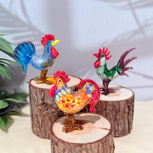 以下が含まれることがあります： カラフルなガラス製の雄鶏の置物3つ。1つは青、もう1つは緑、3つ目はオレンジと黄色です。各雄鶏は赤い鶏冠を持ち、小さな木の輪切りに乗っています。