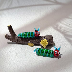 Puede incluir: Dos figuras de oruga de vidrio con caras rojas, antenas azules y cuerpos a rayas verdes y azules. Una oruga descansa sobre un tronco marrón, mientras que la otra está cerca. El tronco tiene detalles amarillos y marrones.
