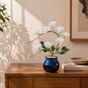 May include: A decorative floral arrangement featuring translucent white flowers and green leaves on thin, arched stems with small clear beads, set in a round, glossy blue and black ceramic pot filled with small white stones.