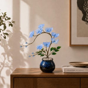 May include: A decorative artificial plant featuring translucent light blue flower blossoms and small blue beads on thin, curved brown stems. The plant has green leaves and is potted in a small, round, dark blue ceramic vase filled with white stones.