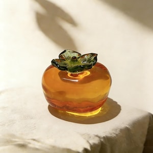 May include: A glass persimmon sculpture with a vibrant orange body and a green, leaf-like top. The sculpture is round and sits on a white surface, casting a shadow. The design is reminiscent of a fruit.
