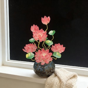 May include: A decorative arrangement of pink lotus flowers in a small, blue ceramic vase. The flowers have translucent petals and green leaves. The vase sits on a white windowsill with a cream-coloured knitted item.