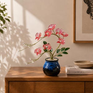 May include: A decorative artificial plant featuring translucent pink flower blossoms with clear crystal centers, green leaves, and thin wire stems. The plant is potted in a small, round, blue and black glazed ceramic vase filled with white pebbles.