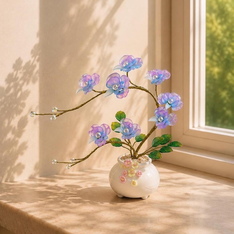 May include: A decorative artificial plant featuring translucent blue and purple orchid-like flowers with clear crystal centers, set in a small white ceramic pot adorned with tiny pastel-colored floral accents and green leaves on thin wire stems.