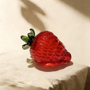 Puede incluir: Una fresa de cristal rojo vibrante con hojas verdes. La fruta translúcida tiene una superficie texturizada que atrapa la luz. Un elemento decorativo único.