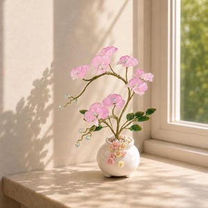 May include: A decorative wire tree sculpture featuring translucent pink flower blossoms and green leaves. The tree is set in a small, round white ceramic pot adorned with tiny pastel-colored floral accents and filled with small white pebbles.