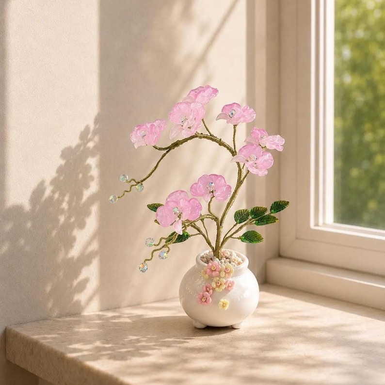May include: A decorative wire tree sculpture featuring translucent pink flower blossoms and green leaves. The tree is set in a small, round white ceramic pot adorned with tiny pastel-colored floral accents and filled with small white pebbles.
