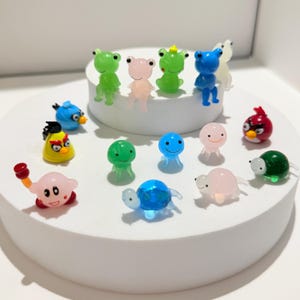 以下が含まれることがあります： カエル、鳥、その他の漫画のキャラクターなど、小さくてカラフルなフィギュアのコレクション。 フィギュアは、緑、ピンク、青、赤などのさまざまな色で、白い円形のプラットフォームに配置されています。