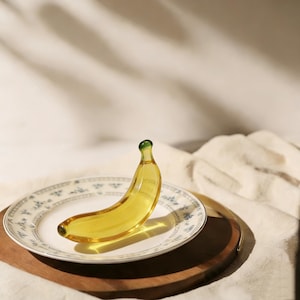 May include: A yellow glass banana figurine with a green tip, resting on a white plate with a blue floral pattern. The plate sits on a wooden tray, with a soft, white fabric in the background.
