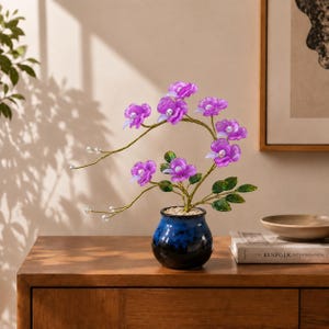 May include: A decorative artificial plant featuring vibrant purple translucent flowers with white centers, arranged on thin gold-toned wire stems with green leaves. The plant is potted in a small, round, glossy blue ceramic vase filled with white pebbles.