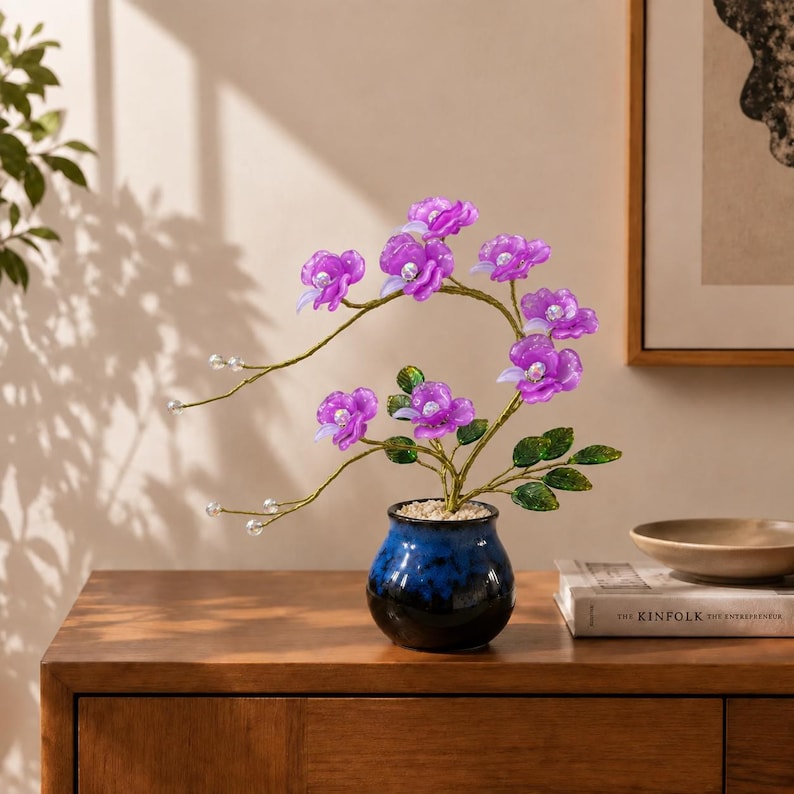 May include: A decorative artificial plant featuring vibrant purple translucent flowers with white centers, arranged on thin gold-toned wire stems with green leaves. The plant is potted in a small, round, glossy blue ceramic vase filled with white pebbles.