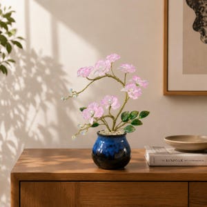May include: A decorative artificial orchid plant with translucent light pink petals and small white centers, featuring green leaves and thin gold-toned wire stems, potted in a small, round, glossy blue and black ceramic vase filled with white pebbles.