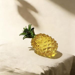 May include: A decorative glass pineapple with a yellow body and green leaves. The pineapple has a textured surface and is resting on a white surface. The image is well-lit, highlighting the details of the glass.