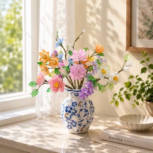 May include: A white ceramic vase with blue floral patterns holds a bouquet of artificial flowers in shades of pink, orange, blue, white, and purple. The flowers feature translucent petals and green leaves on thin stems, creating a vibrant display.