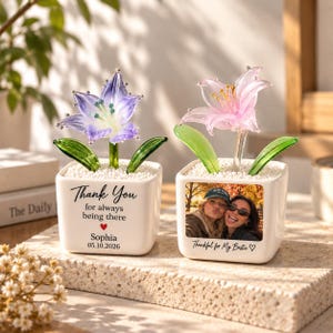 May include: Two square white ceramic planters containing glass flowers. The left planter features a purple glass lily and text reading Thank You for always being there, Sophia, 05.10.2026. The right planter has a pink glass lily and a photo of two people.