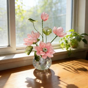 Puede incluir: Un arreglo decorativo de flores de loto rosas y hojas verdes en un jarrón de cristal facetado. Las flores están dispuestas sobre finos tallos verdes. El jarrón está sobre una superficie de madera cerca de una ventana, iluminada por el sol.
