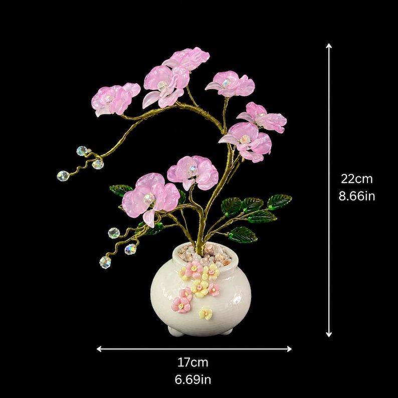 May include: A decorative plant sculpture featuring translucent pink orchid-like flowers on gold-toned wire branches with green leaves and clear beads. The plant sits in a white ceramic pot adorned with small pastel flowers, measuring 8.66 inches high and 6.69 inches wide.
