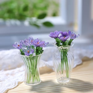 Puede incluir: Dos pequeños jarrones de cristal, cada uno con un ramo de flores moradas con tallos verdes. Los jarrones son transparentes, con un diseño acanalado. Las flores tienen centros naranjas y están dispuestas de forma realista.