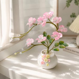May include: A decorative bonsai tree featuring translucent pink flower blossoms with iridescent centers, green leaves, and thin gold wire branches. The tree is planted in a small white ceramic pot adorned with tiny pink and yellow flower accents.